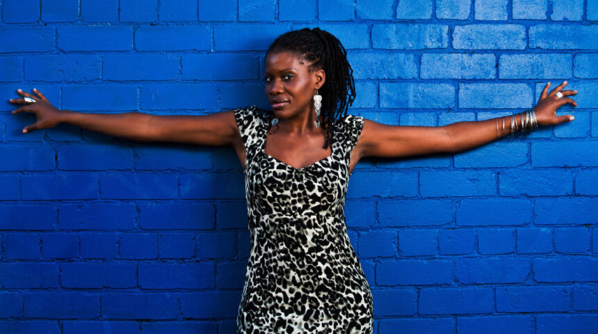 Paula Baxter with arms outstretched against a blue brick wall